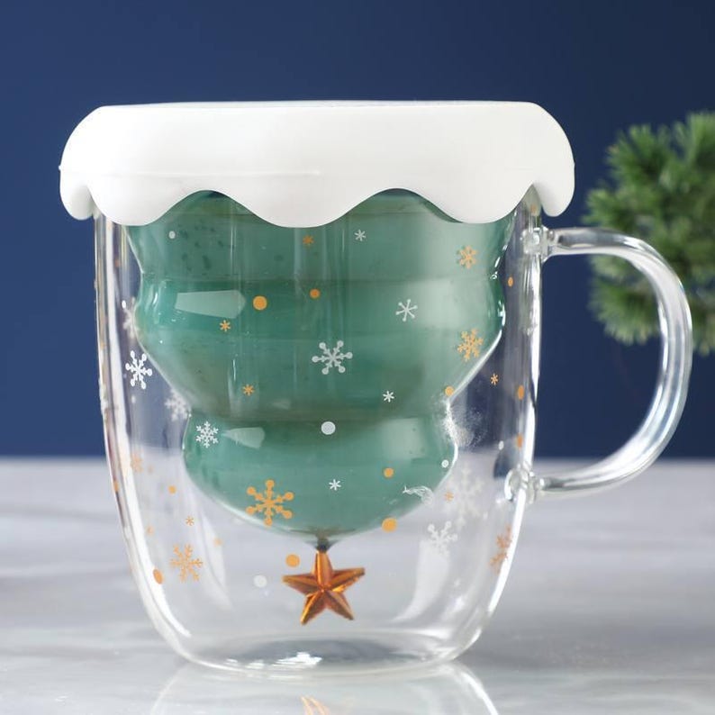 May include: A clear glass mug with a green Christmas tree design inside. The mug has a white, wavy lid and a handle. Gold and white snowflakes and a gold star decorate the exterior. The mug is designed for hot beverages.