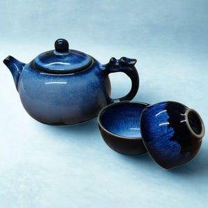 Puede incluir: Una tetera de cerámica azul con tapa a juego y dos tazas pequeñas. La tetera tiene un diseño de asa único y un esmalte azul oscuro que se desvanece a un azul más claro. Las tazas también son azules.