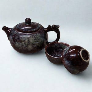 May include: A maroon ceramic tea set comprising a teapot and two teacups. The teapot has a dragon-shaped handle and a matching lid. The teacups are also maroon with a glossy finish. The set is displayed against a white backdrop.