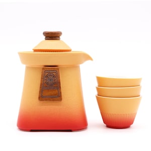 Könnte beinhalten: Ein Keramik-Teeservice mit Farbverlauf. Die Teekanne ist orange mit rotem Boden und einem Deckel und Emblem aus Holz. Drei passende kleine Teetassen befinden sich neben der Teekanne.