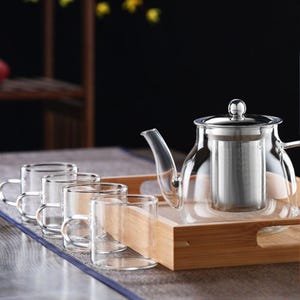 May include: A glass teapot with a stainless steel strainer and lid, along with four clear glass teacups with handles, all arranged on a wooden tray.