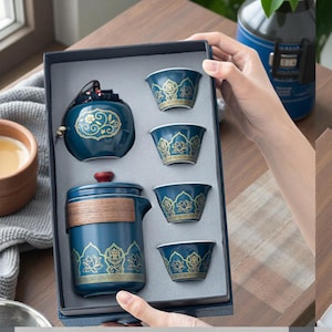 Könnte beinhalten: Ein tealfarbenes Teeservice mit goldenen floralen Mustern, bestehend aus einer Teekanne, einem Teebehälter und vier kleinen Tassen, präsentiert in einer dunkelgrauen Box. Die Teekanne hat einen Holzgriff und einen roten Deckel.
