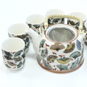 May include: A white ceramic tea set with a teapot and matching cups. The teapot has a woven handle and a built-in strainer. Both the teapot and cups are decorated with colourful fan motifs in shades of green, yellow, and brown.