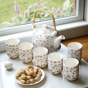 May include: A white ceramic tea set with a floral pattern in brown and grey. The set includes a teapot with a woven handle and six matching cups. A small plate of peanuts and a white sphere are also present.