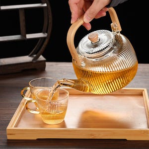 May include: A clear glass teapot with a wooden handle and lid is pouring tea into two matching glass cups. The cups and teapot sit on a bamboo tray. The tea is a golden color.
