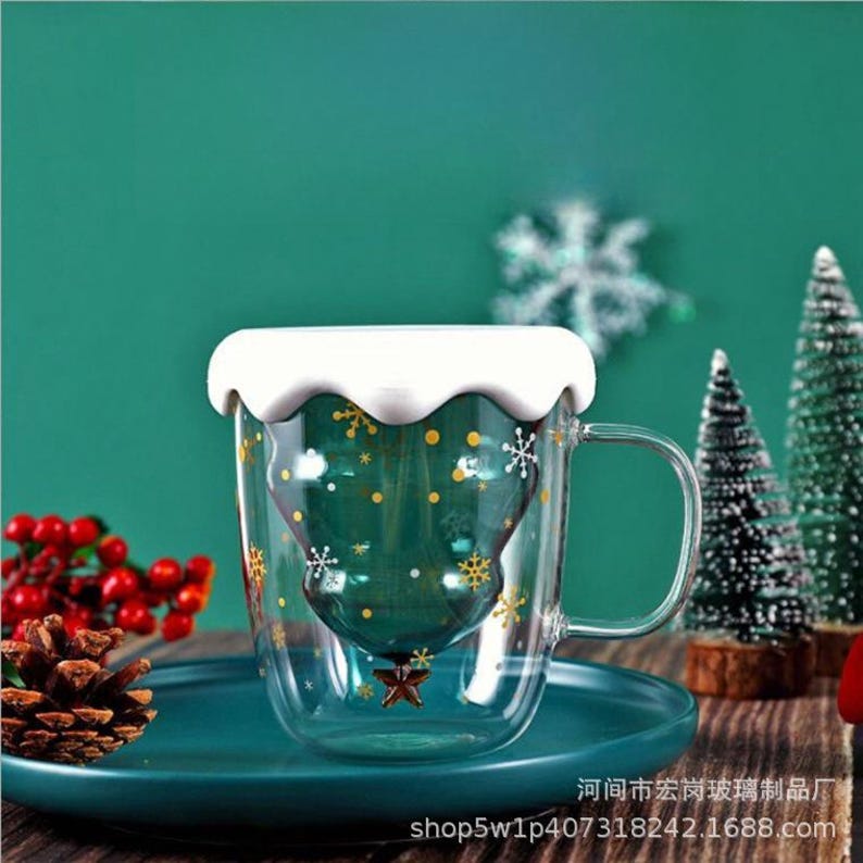 May include: A clear glass mug with a Christmas tree design inside, decorated with snowflakes and gold accents. The mug has a white, wavy rim and a handle. It sits on a teal plate, with a teal background. The text "shop5w1p407318242.1688.com" is visible.