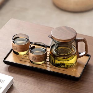 May include: A tea set comprising a glass teapot with a wooden lid and handle, a glass infuser, and two matching glasses with wooden bands. The set is presented on a bamboo tray. A book is in the foreground.