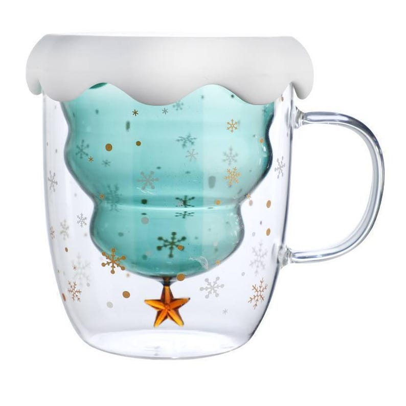 May include: A clear glass mug with a teal-colored, Christmas tree-shaped interior. The mug has a white, wavy rim and a gold star at the bottom. Gold snowflakes and dots decorate the exterior. A clear handle is attached.