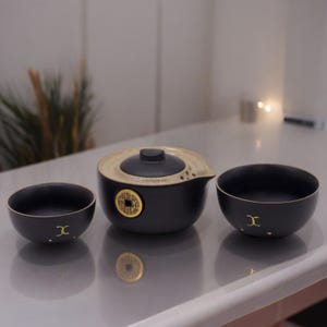 May include: A black ceramic tea set with gold detailing. The set comprises a teapot with a lid, and two matching teacups. The teapot and teacups feature a gold coin design. The tea set is on a reflective surface.