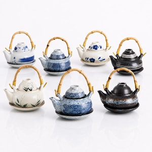 May include: A collection of six ceramic teapots, each featuring a curved bamboo handle and a matching saucer. The teapots display various patterns including blue floral motifs, geometric grids, wave designs, and solid black or speckled finishes.