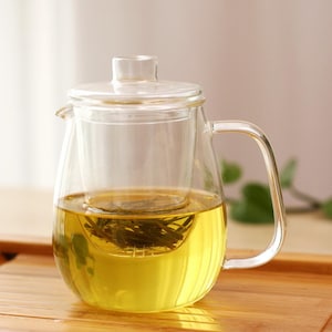 May include: A clear glass teapot with a built-in infuser, filled with yellow tea and loose tea leaves. The teapot has a lid and a handle.