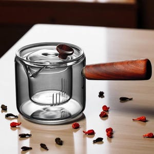 Puede incluir: Una tetera de vidrio transparente con asa y tapa de madera. La tetera tiene un infusor incorporado para preparar té. El asa y el pomo de la tapa son de madera oscura. Hierbas y especias secas están esparcidas alrededor de la tetera.