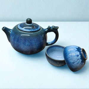 Op de afbeelding: Een blauwe keramische theepot met een deksel en een draakvormige handgreep, samen met twee kleine theekopjes. De theepot en kopjes hebben een glanzende afwerking met een gradiënteffect, van donkerblauw naar lichtere tinten.