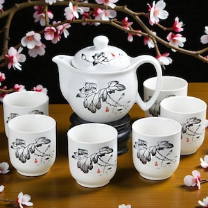 May include: A white ceramic tea set comprising a teapot and six cups. Each item is decorated with a black and grey floral design with red accents. The set is presented on a wooden surface, with flowering branches in the background.