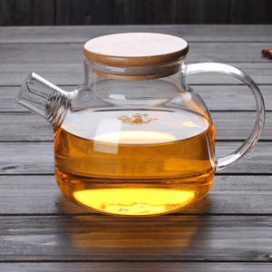 May include: A clear glass teapot with a wooden lid and a handle. The teapot is filled with a light amber liquid.