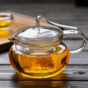 May include: A clear glass teapot with a rounded body and a matching lid. The teapot features a curved handle and a spout, filled with a golden-coloured liquid and a submerged flower. The design includes a unique, elevated glass handle on the lid.
