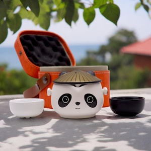 May include: A white panda-themed tea set with a straw hat-shaped lid, black accents, and two small cups. The set includes an orange carrying case with a handle. The tea set is designed for one person.