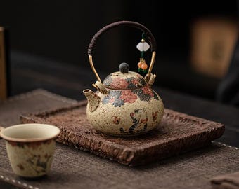 May include: A ceramic teapot with a speckled cream finish, featuring a floral pattern and a cat illustration. It has a dark, wrapped handle with a small decorative charm attached. A matching small cup sits nearby on a textured brown tray.