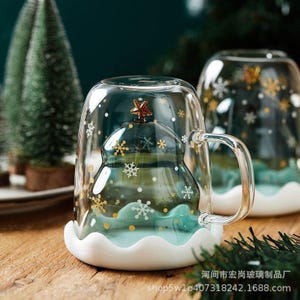 May include: A clear glass mug with a Christmas tree design inside. The tree is green with a gold star on top and sits on a blue base. White snowflakes and gold dots decorate the outside of the mug. The mug has a white scalloped base.