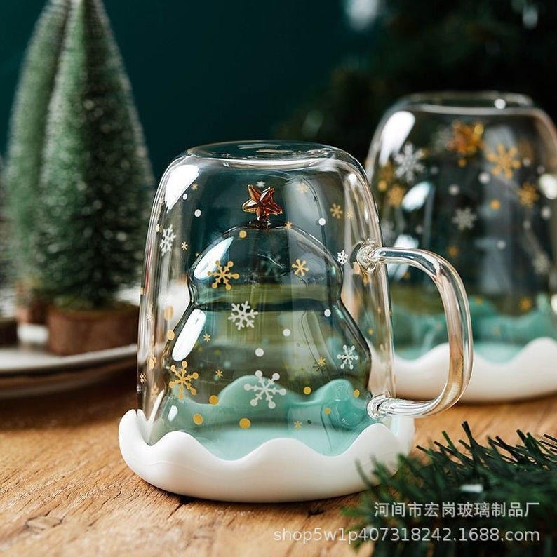 May include: A clear glass mug with a Christmas tree design inside. The tree is green with a gold star on top and sits on a blue base. White snowflakes and gold dots decorate the outside of the mug. The mug has a white scalloped base.