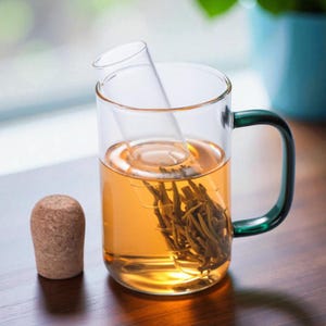 May include: A clear glass tea mug with a green handle and a built-in glass infuser. The mug is filled with amber-coloured tea and tea leaves. A cork stopper sits next to the mug. The mug is designed for steeping loose leaf tea.