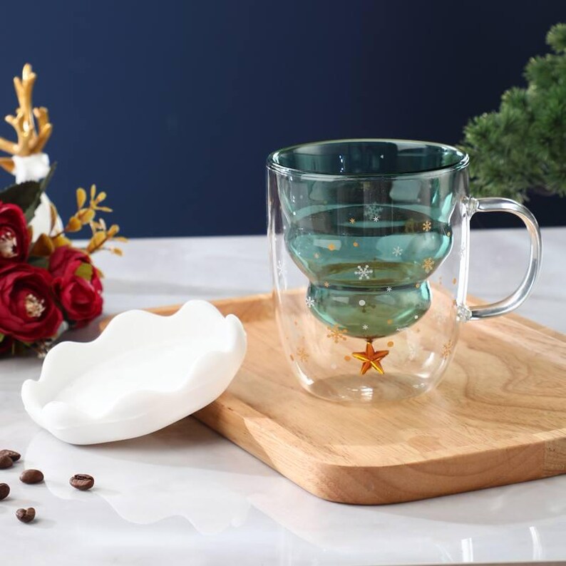 May include: A clear glass mug with a Christmas tree design, featuring a teal interior and snowflake accents. A gold star adorns the base. A white, wavy-edged dish sits beside the mug on a wooden tray. Coffee beans are scattered nearby.