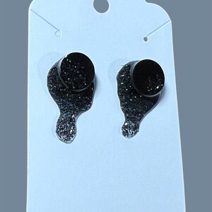 以下が含まれることがあります： 銀色のグリッターが付いた黒いスタッドピアスのペア。ピアスは円形で、下から涙の形がぶら下がっています。