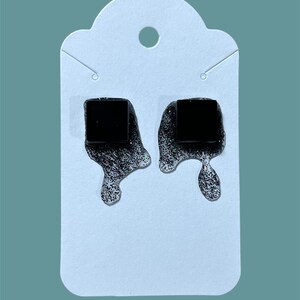 以下が含まれることがあります： キラキラと滴るような効果のある黒い正方形のスタッドピアスのペア。ピアスは、上部に穴が開いた白いカードに表示されています。