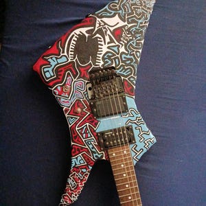 Puede incluir: Una guitarra eléctrica con un diseño abstracto único. El cuerpo presenta una mezcla de patrones rojos, azules y negros sobre un fondo blanco. La guitarra tiene un puente, pastillas y clavijas de afinación negros, con un diapasón marrón.
