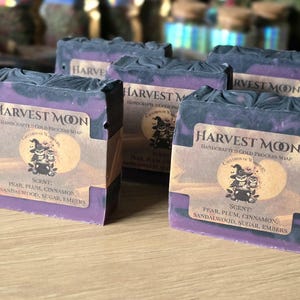 Könnte beinhalten: Mehrere handgemachte Seifenstücke von Harvest Moon. Die Seife hat eine violette Basis mit dunkelgrauen Wirbeln. Jedes Stück hat ein Etikett mit dem Text "Harvest Moon" und der Duftbeschreibung: Birne, Pflaume, Zimt, Sandelholz, Zucker, Glut.