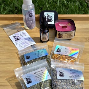May include: A collection of spiritual and ritual items, including a spray bottle, essential oil, candle, incense sticks, and various dried herbs in clear bags. The items are arranged on a wooden tray, with labels indicating scents and uses.