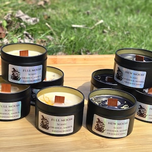 May include: A collection of black tin candles with wooden wicks, labelled "Full Moon" and "New Moon", with scent details. The labels feature a witch graphic. The candles are on a wooden tray.