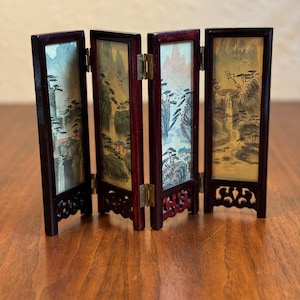 May include: A four-panel folding screen with a dark wood frame and painted scenes of mountains, trees, and waterfalls. The screen is closed and standing on a wooden surface.