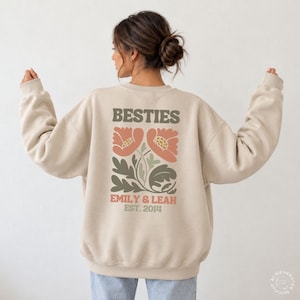 May include: A beige crewneck sweatshirt featuring a back graphic with the text BESTIES, EMILY & LEAH, and EST. 2014. The design includes two large coral-colored flowers with green leaves and stems, rendered in a flat, stylized, and muted color palette.