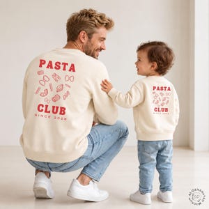 May include: Two cream-colored crewneck sweatshirts featuring red text that reads PASTA CLUB SINCE 2026, accompanied by various red pasta shape illustrations on the back. The sweatshirts are paired with light blue denim jeans and white sneakers.