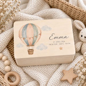 May include: A rectangular wooden keepsake box featuring a watercolor illustration of a hot air balloon in soft blue and peach tones, surrounded by clouds. The text reads Emma, 27. APRIL 2024, 08:36 UHR, 3210 G, 50 CM, and a small star.