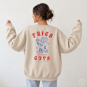 Könnte beinhalten: Ein beigefarbenes Sweatshirt mit Rundhalsausschnitt und einem Grafikdruck auf der Rückseite mit dem roten Schriftzug FRIES BEFORE GUYS sowie dem kleineren Text SINCE 2026. Das Design zeigt eine blaue Cartoon-Illustration einer Pommes-Schachtel.