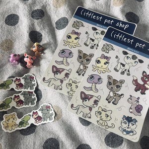 May include: Two sticker sheets and several small animal figurines are displayed on a dotted fabric. The sticker sheets feature various cartoon animals, including cats, dogs, and an octopus. The text "littlest pet shop" is printed on the sticker sheets.