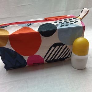 Op de afbeelding: Een witte toilettas met een kleurrijk abstract patroon van cirkels en strepen. De tas heeft een ritssluiting en een gele handgreep. Een witte en gele roll-on deodorant ligt naast de tas.