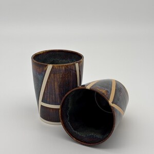 May include: Two ceramic tumblers featuring a dark brown and blue reactive glaze with a glossy finish. Each cup is decorated with a series of intersecting cream-colored lines that form geometric shapes across the exterior surface of the vessels.