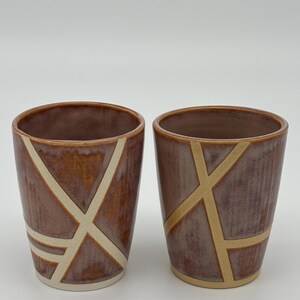 Op de afbeelding: Twee keramische bekers met een glanzend, roodbruin geglazuurd oppervlak. Elke beker is versierd met een reeks kruisende, ongeglazuurde cr&egrave;mekleurige lijnen die geometrische patronen vormen op de buitenkant van de drinkbekers.