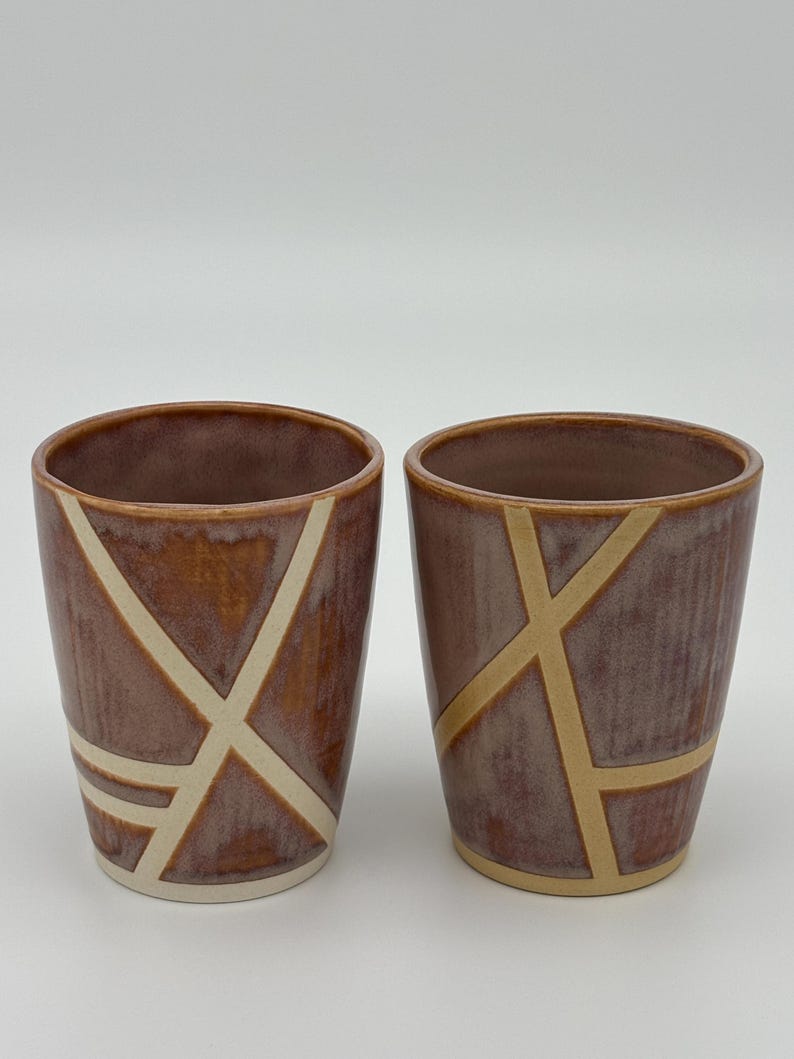 Op de afbeelding: Twee keramische bekers met een glanzend, roodbruin geglazuurd oppervlak. Elke beker is versierd met een reeks kruisende, ongeglazuurde cr&egrave;mekleurige lijnen die geometrische patronen vormen op de buitenkant van de drinkbekers.