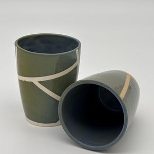 Op de afbeelding: Twee keramische bekers met een donker olijfgroene buitenkant en een diepblauwe binnenkant. De rechtopstaande beker heeft witte lineaire geometrische patronen, terwijl de gekantelde beker een enkele verticale beige lijn aan de zijkant toont.