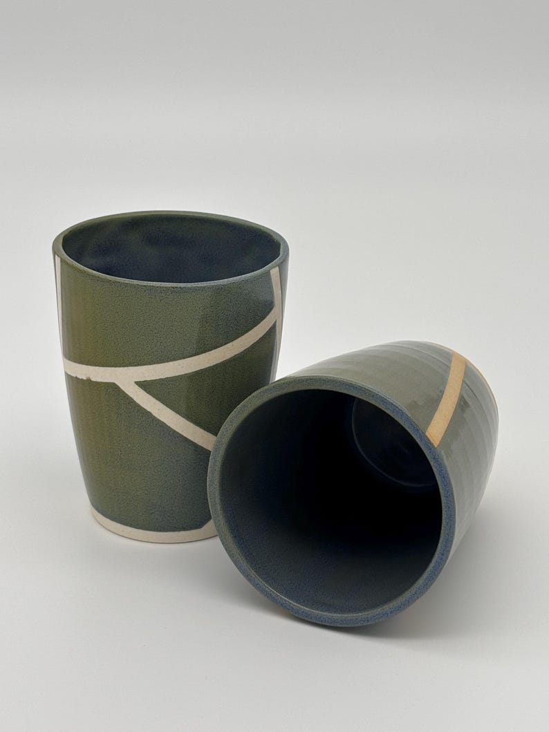 Op de afbeelding: Twee keramische bekers met een donker olijfgroene buitenkant en een diepblauwe binnenkant. De rechtopstaande beker heeft witte lineaire geometrische patronen, terwijl de gekantelde beker een enkele verticale beige lijn aan de zijkant toont.