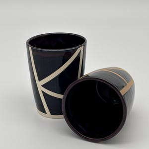 Op de afbeelding: Twee donker marineblauwe keramische bekers met een geometrisch patroon van dunne, lichtbeige lijnen. De ene beker staat rechtop, terwijl de andere op zijn kant ligt, waardoor de gladde, donkere binnenkant en de onderkant zichtbaar zijn.