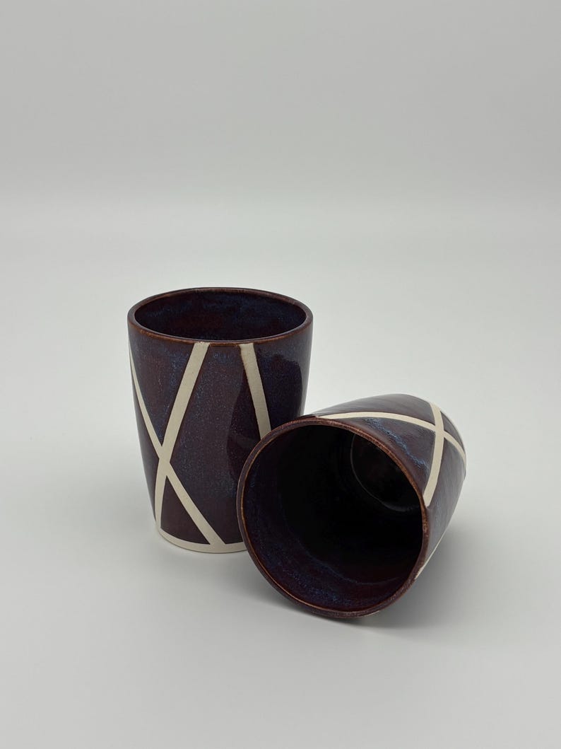 Op de afbeelding: Twee keramische bekers met een glanzend dieppaarse glazuurlaag en een contrasterend wit geometrisch lijnpatroon. De ene beker staat rechtop terwijl de andere op zijn kant ligt, wat de gladde binnenkant en het ontwerp van de buitenkant laat zien.