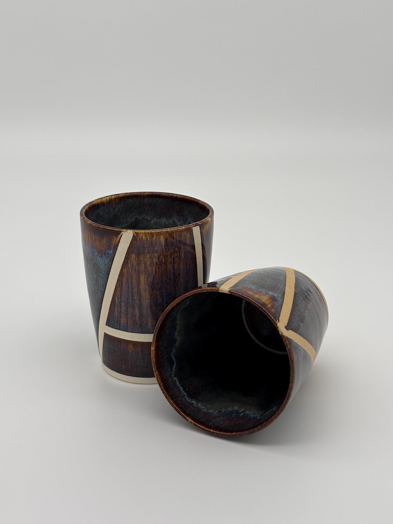 Op de afbeelding: Twee keramische bekers met een donkerbruin en blauw reactief glazuur met een glanzende afwerking. Elke beker is versierd met een reeks kruisende cr&egrave;mekleurige lijnen die geometrische vormen vormen op het buitenoppervlak van de vaten.