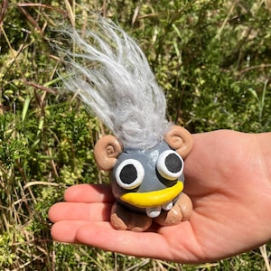 May include: A gray troll figurine with a white fuzzy hair, brown ears, and a yellow beak. The troll has large white eyes with black pupils and a single white tooth.
