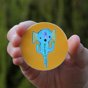 May include: A blue and green cartoon creature with a flower on its head is printed on a round yellow sticker.