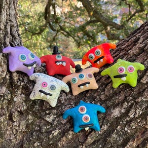 May include: A collection of colorful, handmade monster figurines. Each monster is a different color, including purple, red, orange, green, blue, and tan. They have multiple eyes, small hats, and smiling mouths. The figurines are displayed on a tree trunk.